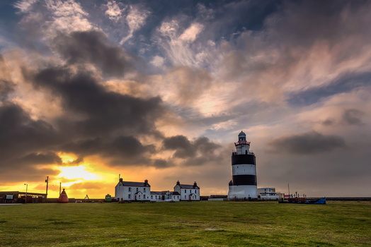 View of Hook Lighthouse with its iconic black and white stripes and adjacent white cottages under a cloudy sunset sky with golden light in County Wexford Ireland. View of Hook Lighthouse with its iconic black and white stripes and adjacent white cottages under a cloudy sunset sky with golden light in County Wexford Ireland.