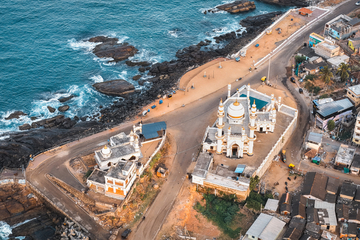 Aerial view of the Vizhinjam Mosque situated along the rocky coastline with blue sea waves nearby roads and surrounding urban structures in Kovalam Keralam India. Aerial view of the Vizhinjam Mosque situated along the rocky coastline with blue sea waves nearby roads and surrounding urban structures in Kovalam Keralam India.