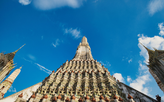 View of the ornate central prang of Wat Arun temple decorated with colorful porcelain mosaics under a bright blue sky in Wat Arun Bangkok Thailand.