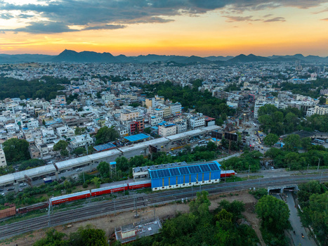 Aerial view of a train station with a red train parked at the platform surrounded by urban buildings lush trees and distant hills under a vibrant sunset sky in Udaipur Rajasthan India.