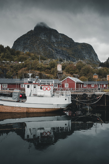 Nusfjord Nordland Norway 07 September 2022 View of Nusfjord village with a white boat moored at the pier reflecting in calm water against dramatic mountains and red buildings. Nusfjord Nordland Norway 07 September 2022 View of Nusfjord village with a white boat moored at the pier reflecting in calm water against dramatic mountains and red buildings.