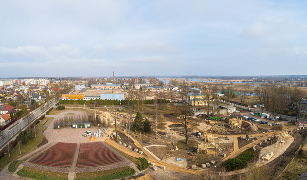 Aerial view of a park under construction featuring wooden play structures paved paths and benches with nearby residential buildings and a river in Latvia. Aerial view of a park under construction featuring wooden play structures paved paths and benches with nearby residential buildings and a river in Latvia.