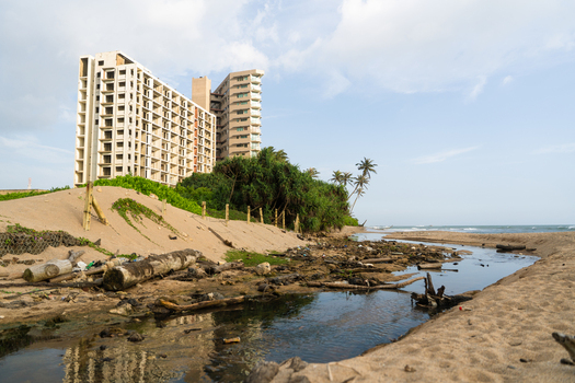 View of a modern high rise building under construction near a sandy beach with a debris filled stream and lush palm trees in Galle Southern Province Sri Lanka.