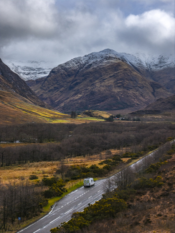 Aerial view of a white campervan driving on a winding road through a valley with snow capped mountains under a cloudy sky Scotland United Kingdom. Aerial view of a white campervan driving on a winding road through a valley with snow capped mountains under a cloudy sky Scotland United Kingdom.