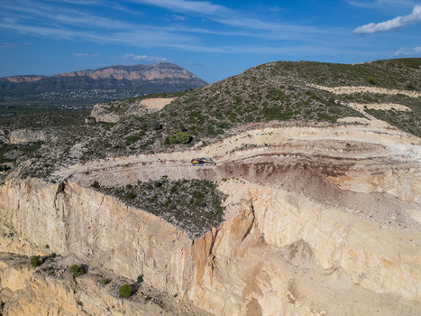 Aerial view of an active stone quarry with an excavator on a terraced hillside surrounded by rugged landscape under a clear blue sky in Teulada Valencian Community Spain.