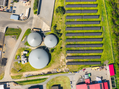 Aerial view of a solar farm with rows of photovoltaic panels adjacent to large industrial biogas storage tanks and facility buildings in Brno South Moravian Region Czechia.