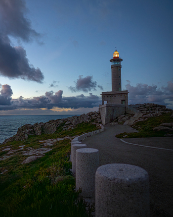 View of Faro de Punta Nariga lighthouse with its lantern illuminated on rocky cliffs under a twilight sky in Malpica A Coruna Spain. View of Faro de Punta Nariga lighthouse with its lantern illuminated on rocky cliffs under a twilight sky in Malpica A Coruna Spain.