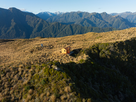Aerial view of a solitary cabin glows warmly amidst the rugged terrain contrasting with the distant snow capped peaks Lake Kaniere West Coast Region New Zealand. Aerial view of a solitary cabin glows warmly amidst the rugged terrain contrasting with the distant snow capped peaks Lake Kaniere West Coast Region New Zealand.