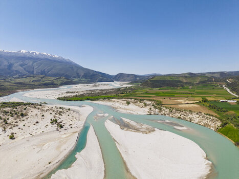 Aerial view of Vjosa Wild River National Park with turquoise river white gravel riverbeds and green agricultural fields under a clear sky in Permet Gjirokaster County Albania. Aerial view of Vjosa Wild River National Park with turquoise river white gravel riverbeds and green agricultural fields under a clear sky in Permet Gjirokaster County Albania.