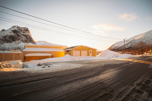 View of golden light kissing the snow covered road and buildings with majestic mountains rising in the background Indregård Troms Norway. View of golden light kissing the snow covered road and buildings with majestic mountains rising in the background Indregård Troms Norway.