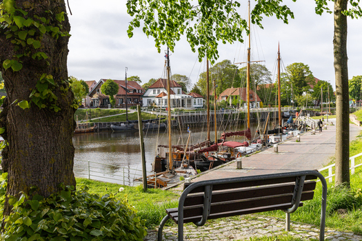 View of Hafen Harlesiel with traditional sailing boats moored along the canal featuring a wooden bench and lush green trees in the foreground Wittmund Lower Saxony Germany. View of Hafen Harlesiel with traditional sailing boats moored along the canal featuring a wooden bench and lush green trees in the foreground Wittmund Lower Saxony Germany.
