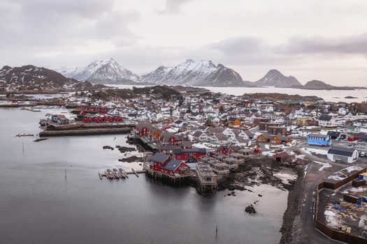 Aerial view of red cabins clinging to the rocky shore contrast with the snow dusted village and the jagged peaks in the distance Ballstad Nordland Norway. Aerial view of red cabins clinging to the rocky shore contrast with the snow dusted village and the jagged peaks in the distance Ballstad Nordland Norway.