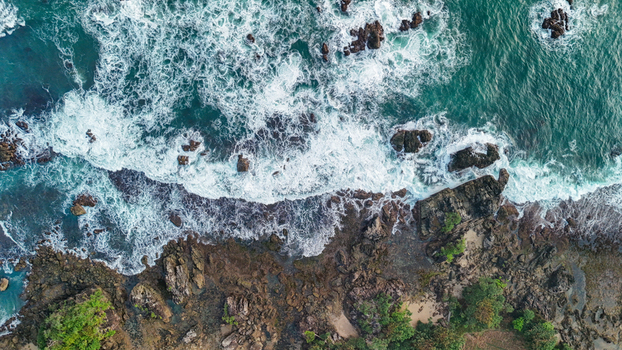 Aerial view of waves crashing against a rocky coastline with lush green vegetation in Indonesia. Aerial view of waves crashing against a rocky coastline with lush green vegetation in Indonesia.