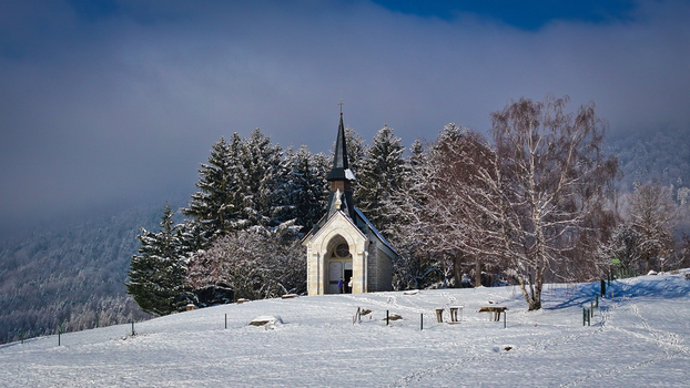 Aerial view of the Chapelle de Riantmont on a snow covered hill surrounded by frosted pine trees and misty mountains in Vesancy Auvergne Rhone Alpes France. Aerial view of the Chapelle de Riantmont on a snow covered hill surrounded by frosted pine trees and misty mountains in Vesancy Auvergne Rhone Alpes France.