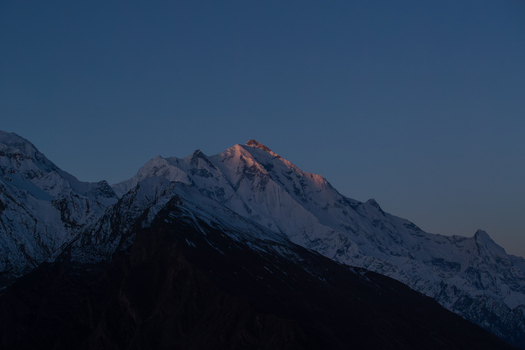 View of the snow capped peak of Rakaposhi mountain glowing with warm light at sunset against a deep blue sky in Hunza Nagar Gilgit Pakistan. View of the snow capped peak of Rakaposhi mountain glowing with warm light at sunset against a deep blue sky in Hunza Nagar Gilgit Pakistan.