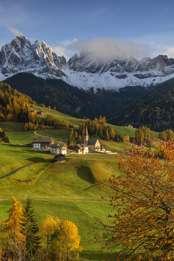 View of Church of St. John nestled in green meadows with the snow covered Odle mountain peaks and autumn foliage Funes Trentino Alto Adige Italy.