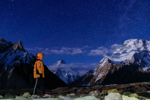 View of a person in an orange jacket looking at the snow capped K2 mountain under a starry night sky in Skardu Gilgit Pakistan.