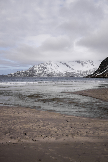 View of a sandy beach with a small stream flowing into the sea under snow capped mountains and a cloudy sky in Kvaloya Troms Norway. View of a sandy beach with a small stream flowing into the sea under snow capped mountains and a cloudy sky in Kvaloya Troms Norway.