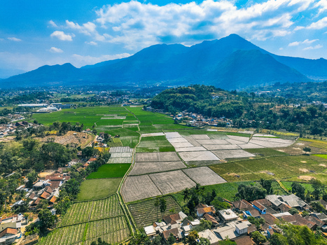 Aerial view of lush green rice fields and a small village nestled at the foot of mountains under a bright blue sky in Garut West Java Indonesia. Aerial view of lush green rice fields and a small village nestled at the foot of mountains under a bright blue sky in Garut West Java Indonesia.