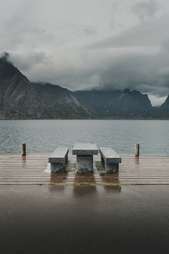 View of a stone table and benches on a wooden pier overlooking the water with dramatic mountains under a cloudy sky in Sakrisoya Moskenes Nordland Norway.
