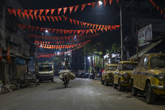 Kolkata India 06 December 2024 View of a cyclist carrying a large load passes beneath vibrant red pennants strung across a darkened street lined with parked yellow taxis. Kolkata India 06 December 2024 View of a cyclist carrying a large load passes beneath vibrant red pennants strung across a darkened street lined with parked yellow taxis.
