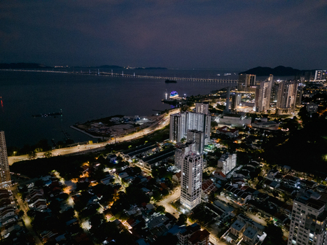 Aerial view of the illuminated cityscape of George Town at night featuring high rise buildings coastal roads and the distant Penang Bridge in George Town Penang Malaysia. Aerial view of the illuminated cityscape of George Town at night featuring high rise buildings coastal roads and the distant Penang Bridge in George Town Penang Malaysia.