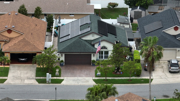 Aerial view of a suburban house with solar panels on the roof and an American flag in the front yard surrounded by green lawns and palm trees in Orlando Florida United States. Aerial view of a suburban house with solar panels on the roof and an American flag in the front yard surrounded by green lawns and palm trees in Orlando Florida United States.