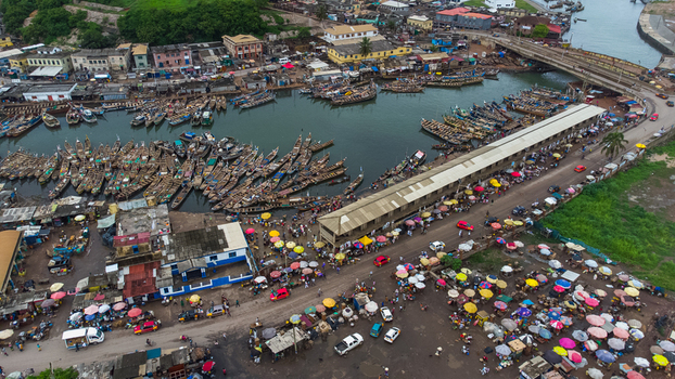 Aerial view of Elmina fishing harbour with numerous wooden boats docked in the water a bustling market with colorful umbrellas along the shore and a bridge in Elmina Central Region Ghana. Aerial view of Elmina fishing harbour with numerous wooden boats docked in the water a bustling market with colorful umbrellas along the shore and a bridge in Elmina Central Region Ghana.