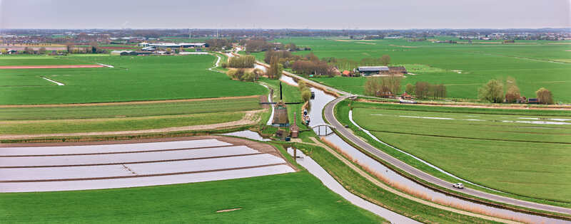 Aerial view of the historic De Kaagmolen windmill standing beside a winding canal surrounded by vast green agricultural fields and a rural road Spanbroek North Holland Netherlands. Aerial view of the historic De Kaagmolen windmill standing beside a winding canal surrounded by vast green agricultural fields and a rural road Spanbroek North Holland Netherlands.