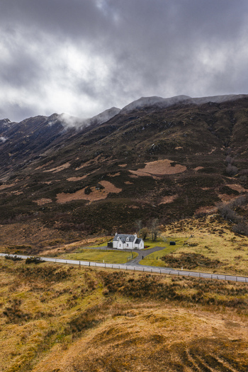 Aerial view of a lonely white cottage at the foot of rugged mountains under a moody overcast sky in Scotland United Kingdom. Aerial view of a lonely white cottage at the foot of rugged mountains under a moody overcast sky in Scotland United Kingdom.