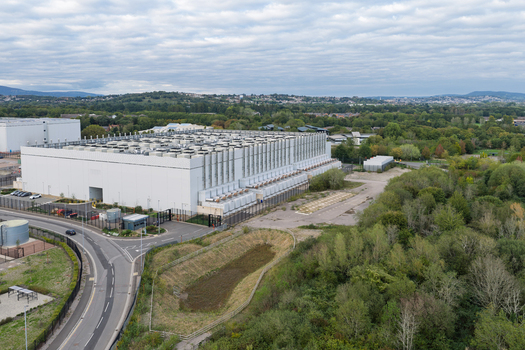 Aerial view of a large modern data center facility with cooling units on the roof surrounded by green trees a road and a parking area in the United Kingdom.