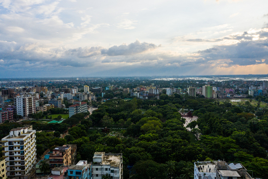 Aerial view of the dense urban landscape of Dhaka featuring residential buildings lush green parks and scattered trees under a cloudy sunset sky in Dhaka Dhaka Division Bangladesh.