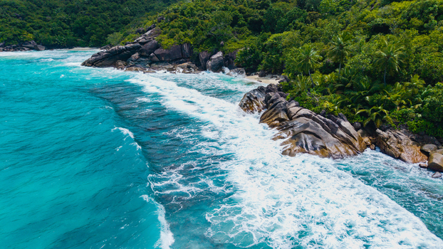 Aerial view of Anse Soleil Beachcomber Hotel surrounded by lush tropical vegetation and turquoise ocean waves crashing against granite rocks in Mahe Baie Lazare Seychelles.