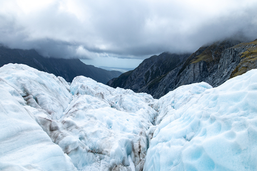 View of the icy glacier s blue and white hues contrasting with the dark jagged mountains under a cloudy sky Franz Josef Glacier West Coast Region New Zealand. View of the icy glacier s blue and white hues contrasting with the dark jagged mountains under a cloudy sky Franz Josef Glacier West Coast Region New Zealand.
