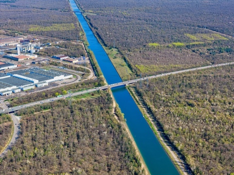 Aerial view of a tranquil canal cutting through a dense forest reflecting the sky s azure hue juxtaposed with the industrial buildings Sausheim Grand Est France.