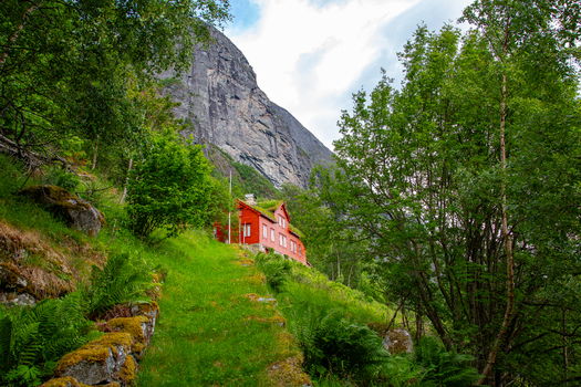 View of Smage mountain farm a traditional red wooden house with a grass roof nestled on a lush green hillside beneath a towering rocky cliff in Geiranger More og Romsdal Norway.