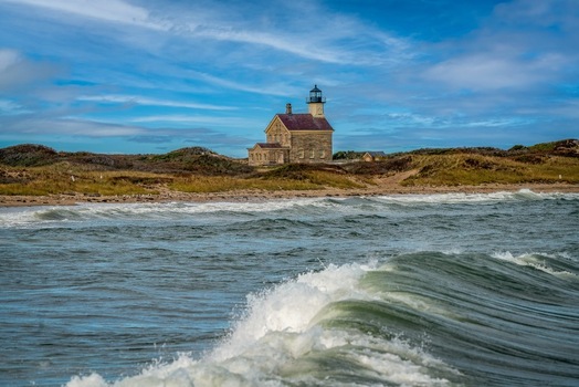 View of the historic Block Island North Light on grassy dunes above the beach with crashing waves in the foreground under a blue sky in New Shoreham Rhode Island United States.