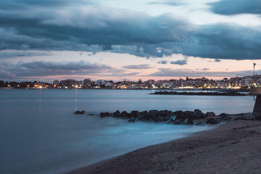 View of the tranquil beach with the city lights glowing softly under a dramatic sky with dark clouds Taormina Sicily Italy. View of the tranquil beach with the city lights glowing softly under a dramatic sky with dark clouds Taormina Sicily Italy.