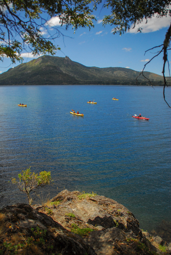 View of kayakers paddling on the blue waters of Nahuel Huapi Lake with distant mountains under a blue sky with light clouds San Carlos de Bariloche Rio Negro Argentina. View of kayakers paddling on the blue waters of Nahuel Huapi Lake with distant mountains under a blue sky with light clouds San Carlos de Bariloche Rio Negro Argentina.