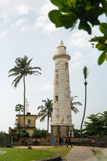 View of the historic Galle Lighthouse surrounded by tall palm trees and stone fortifications with people walking on a path in Galle Southern Province Sri Lanka.