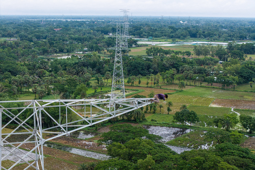 Trishal Upazila Bangladesh 13 May 2026 Aerial view of a worker sitting on a high voltage electricity pylon overlooking a lush green rural landscape with fields and trees. Trishal Upazila Bangladesh 13 May 2026 Aerial view of a worker sitting on a high voltage electricity pylon overlooking a lush green rural landscape with fields and trees.