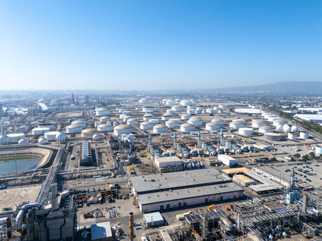 Aerial view of an industrial oil refinery with large white storage tanks complex piping and processing facilities under a clear blue sky in Carson California United States.