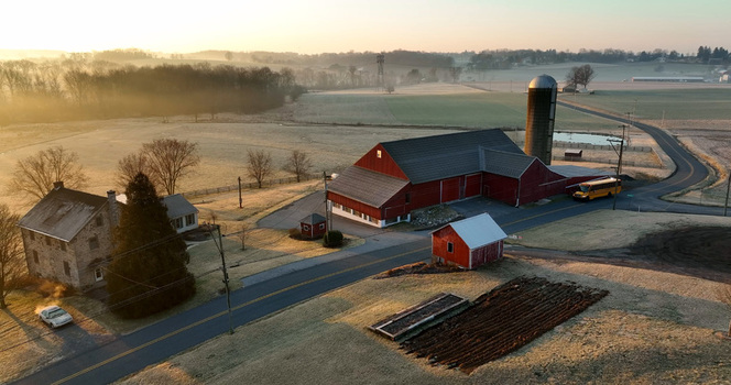 Aerial view of Penryn farmland featuring a red barn with a silo stone farmhouse and school bus on a rural road during a misty sunrise in Penryn Pennsylvania United States.