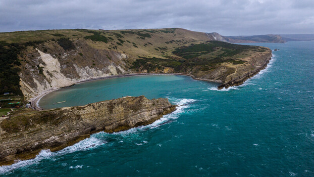 Aerial view of Lulworth Cove featuring turquoise waters dramatic limestone cliffs and rolling grassy hills along the coastline in Wareham England United Kingdom. Aerial view of Lulworth Cove featuring turquoise waters dramatic limestone cliffs and rolling grassy hills along the coastline in Wareham England United Kingdom.