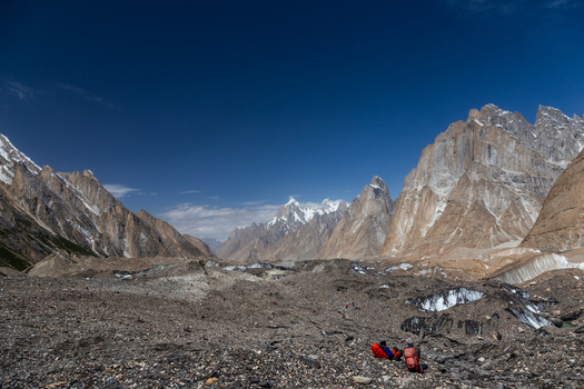 View of two hikers resting on the rocky moraine of the Baltoro Glacier surrounded by towering snow capped peaks under a clear blue sky in Baltoro Gilgit Baltistan Pakistan.