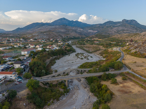 Aerial view of the historic Mesi Bridge spanning the Kir River surrounded by rural landscape stone buildings and mountains in Boks Qarku i Shkodres Albania.