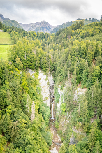 Aerial view of Grandvillard Waterfall cascading down a steep rocky cliff surrounded by lush green forest and alpine mountains under a cloudy sky Grandvillard Fribourg Switzerland.