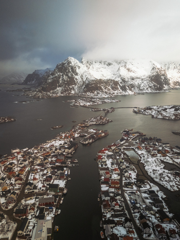 Aerial view of snow dusted rooftops nestled along the rugged coastline contrasting with the stark snow capped mountains fading into the misty horizon Henningsv&aelig;r Nordland Norway.