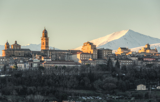 View of the historic town of Macerata with its bell tower and stone buildings illuminated by golden hour light set against a backdrop of snow capped mountains in Macerata Marche Italy. View of the historic town of Macerata with its bell tower and stone buildings illuminated by golden hour light set against a backdrop of snow capped mountains in Macerata Marche Italy.