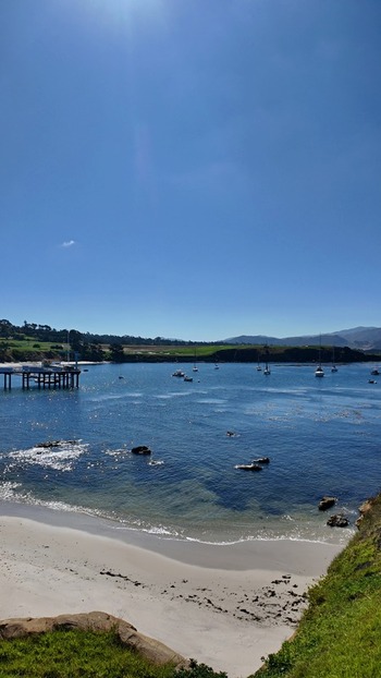 View of Stillwater Cove along 17 Mile Drive with a wooden pier anchored sailboats sandy beach and clear blue sky in Del Monte Forest California United States. View of Stillwater Cove along 17 Mile Drive with a wooden pier anchored sailboats sandy beach and clear blue sky in Del Monte Forest California United States.